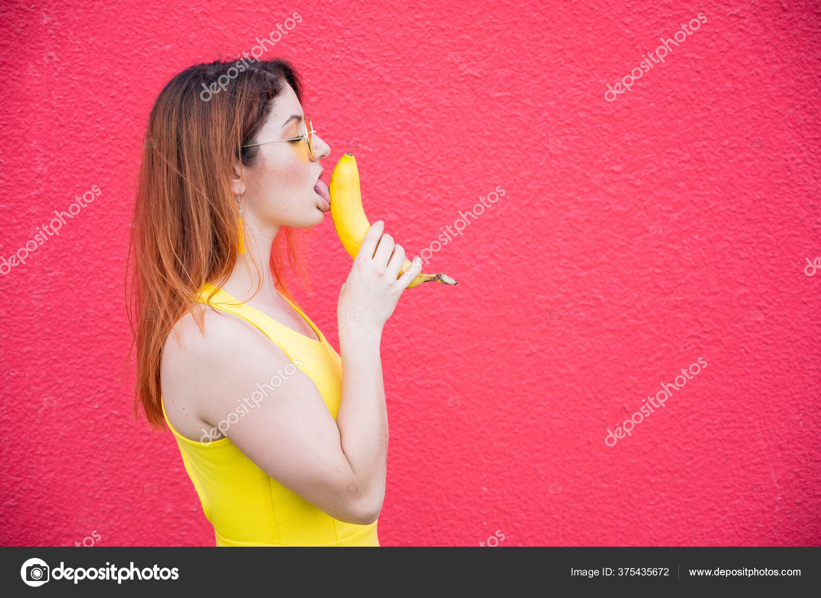 Woman in a yellow dress and glasses erotically licks a banana on a red background