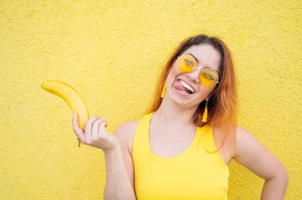 Belle femme aux cheveux roux en robe jaune et lunettes de soleil montre la langue et tient une banane. Portrait d'une fille ludique fantasmant avec des fruits mûrs. Dame avec un sourire éblouissant parfait. — Photo