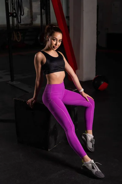 Hermosa joven caucásica posando en un gimnasio oscuro. Una mujer en ropa deportiva está descansando de un entrenamiento circular en una caja. Una ruptura entre conjuntos . —  Fotos de Stock