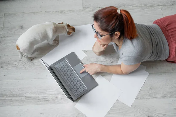 Une femme s'allonge sur le ventre par terre et tape sur un ordinateur portable. Fille pigiste travaille à distance de la maison avec un chiot. Jack Russell Terrier à côté du propriétaire dans une position confortable . — Photo