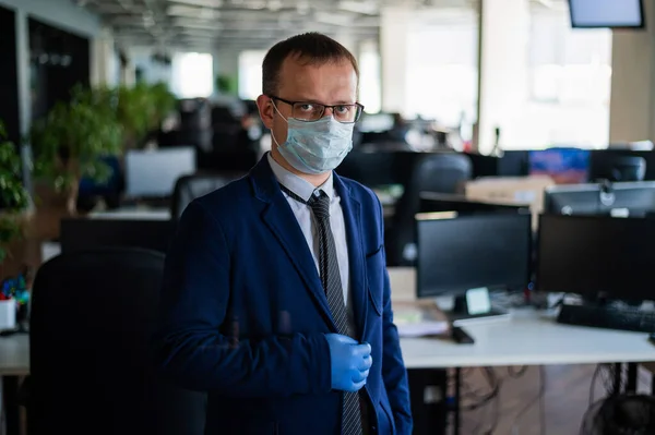 Serious man in a business suit and glasses in an empty office during quarantine. Male manager in a medical mask at the workplace. Social distance and workspace disinfection.