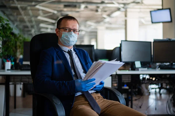 A man in a business suit and medical mask reads a paper report in an empty open space office. Social distance and isolation of employees. Urgent work during quarantine.