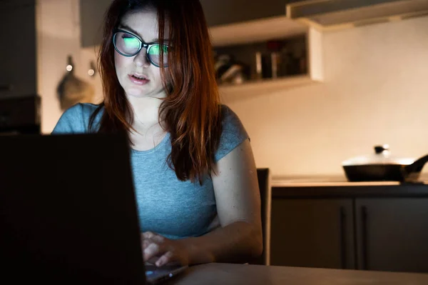 Femme blanche portant des lunettes à un ordinateur sans fil la nuit dans la cuisine. Une fille fait des heures sup 'au bureau. Freelance tape sur un ordinateur portable . — Photo