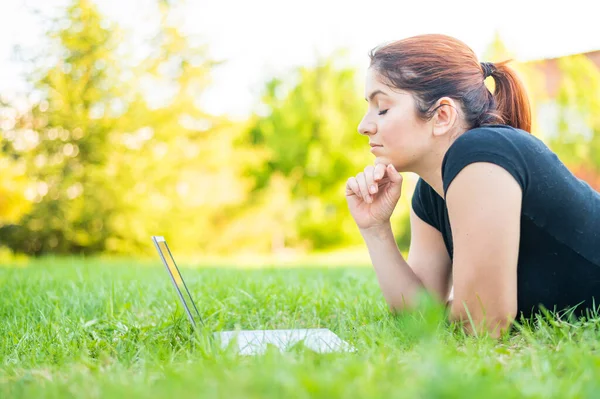 Uma mulher deita-se de barriga em um parque e digita em um laptop. Freelancer feminino trabalha remotamente ao ar livre. Menina está estudando em um computador no gramado . — Fotografia de Stock