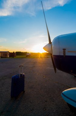 Mavi bir bavul ve park edilmiş küçük özel bir jet. Gün batımında hava taksisi için pervaneli dört kat uçak. Seyahat konsepti. VIP kişiler için uçaklar.