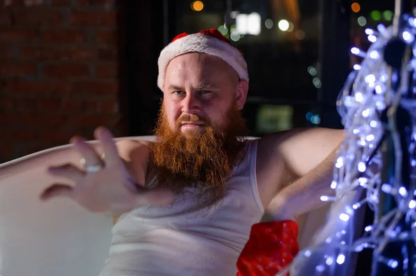 Vicces kép egy gonosz télapóról, aki karácsonyi fényekkel fürdik. Egy férfi vörös szakállal, télapó sapkában és alsóneműben egy koszorút tart a kezében.. — Stock Fotó