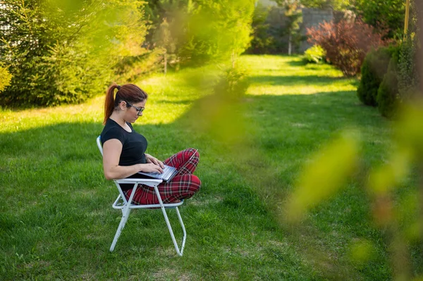 Uma mulher com óculos senta-se em uma cadeira dobrável com as pernas cruzadas em uma posição de lótus e digitando em um laptop. Freelancer feminino trabalha remotamente no jardim da casa de campo durante a quarentena . — Fotografia de Stock