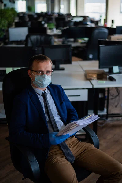 A man in a business suit and medical mask reads a paper report in an empty open space office. Social distance and isolation of employees. Urgent work during quarantine.
