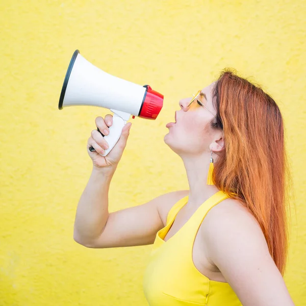 Eine Frau im Kleid mit Brille und Ohrringen steht im Profil vor gelbem Hintergrund und ruft in ein Megafon. Porträt eines Mädchens mit einem Lautsprecher. Lady schreit ein Schallverstärkungsgerät an. — Stockfoto