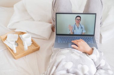 Bir kadın yatağa uzanıyor ve bir terapistin soğuk algınlığı tedavisi hakkında tavsiyelerini dinliyor. Laptopta tıbbi nakli izleyen bir kız. Doktor çevrimiçi danışmanlık yapıyor..