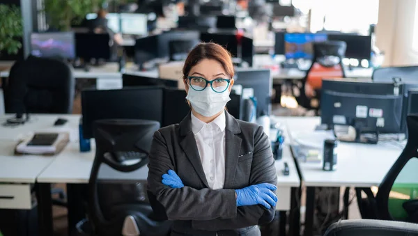 Business woman stands with arms crossed in the office. Female manager in a medical mask and gloves. Work in a coronavirus epidemic. Ways to prevent the spread of infection. Workplace disinfection.