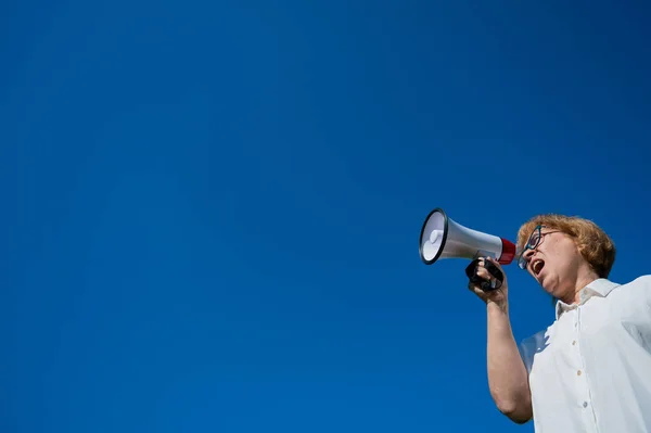 Wütende Seniorin mit Brille, die draußen in ein Megafon ruft. Genervter Rentner schreit in ein Gerät, das Geräusche vor blauem Himmel verstärkt. Lautsprecher in Frauenhand. — Stockfoto