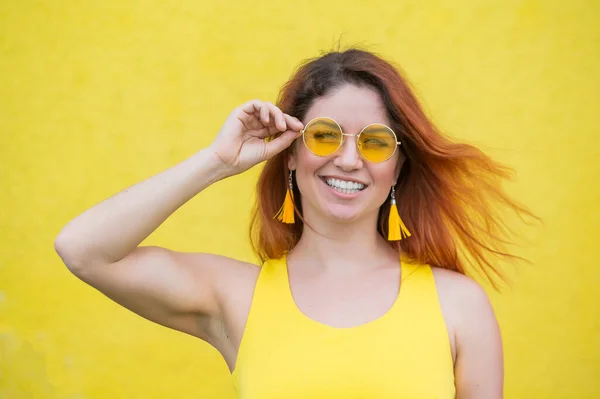 Porträtt av en lycklig kvinna i solglasögon på en gul bakgrund. Rödhårig bekymmerslös flicka i klänning och örhängen tofsar av samma färg. Lady med ett oklanderligt bländande leende. Sommarfoto. — Stockfoto