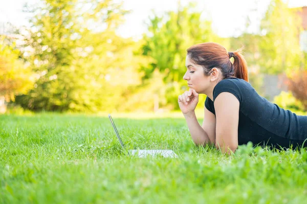 一个女人躺在公园里，在笔记本电脑上打字。女自由职业者在户外远程工作。女孩正在草坪上的电脑上学习. — 图库照片