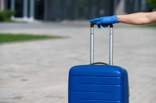 Close-up van een vrouwelijke hand in een handschoen op bagage. Een gezichtloze vrouw houdt een grote blauwe tas bij de uitschuifbare handgreep op straat. Reisconcept tijdens een virusuitbraak. Persoonlijke hygiëne. — Stockfoto