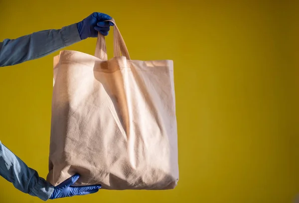 Sending eco-friendly reusable cotton bag on a yellow background. The courier holds a canvas fabric bag without plastic packaging on a yellow background. Safe delivery to the epidemic. — Stock Photo, Image