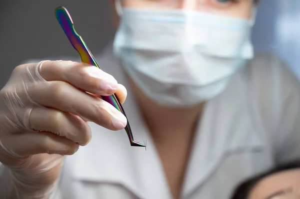 Close-up of tweezers with a bunch of artificial eyelashes in the hands of a leshmaker. The master in eyelash extensions in a mask and gloves holds a sterile instrument.