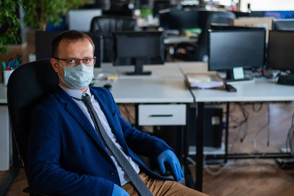 Serious man in a business suit and glasses in an empty office during quarantine. Male manager in a medical mask at the workplace. Social distance and workspace disinfection.