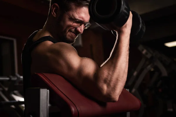 Un bel homme avec des lunettes faisant un exercice pour les biceps avec un haltère. Il s'occupe de musculation. Entraîneur dans le gymnase avec les bras musclés . — Photo