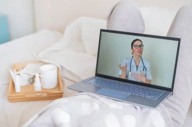 Karantina sırasında bir kadın doktoruyla iletişim kurmak için dizüstü bilgisayar kullanıyor. Kız sırt üstü uzanıyor ve tıbbi video blogu izliyor. Doktor çevrimiçi danışmanlık yapıyor..