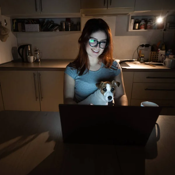 A smiling woman with glasses sits in the dark at a wireless computer in the kitchen with a puppy of Jack Russell Terrier on her knees. Girl with her little dog watching a movie on a laptop at home.