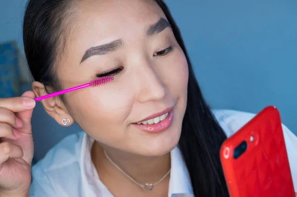 Mulher asiática bonita penteando suas extensões de pestanas com um pincel rosa. Retrato de uma menina tomando uma selfie em um salão de beleza. Procedimento de extensão das pestanas artificiais . — Fotografia de Stock