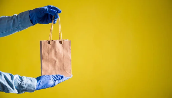 Female courier hand in medical latex gloves holds a small paper bag. Safe delivery during the spread of coronavirus. Ecofriendly packaging. Online shopping. — Stock Photo, Image