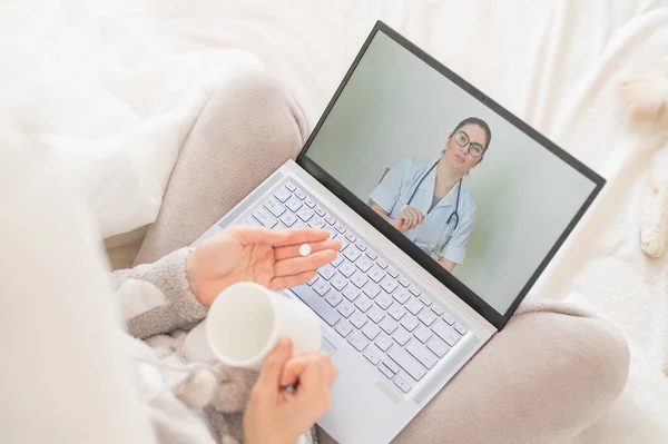 Невпізнавана жінка п'є таблетки і дивиться медичний відео блог на ноутбуці. Жінка-лікар проводить дистанційну консультацію з хворим пацієнтом. Дівчина з грипом на хворій відпустці . — стокове фото