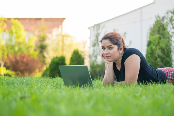 Uma mulher se deita de barriga no gramado da casa de campo e usa em um laptop. Uma freelancer focada trabalha remotamente ao ar livre. A menina está estudando no computador . — Fotografia de Stock