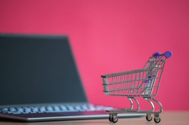Mini alışveriş arabası pembe arka planda dizüstü bilgisayarın önünde duruyor. İnternetten alışveriş konsepti. Siyah bilgisayar ekranı. İnternetten uzaktan satın alınan ürünler falan. Kimse yok..