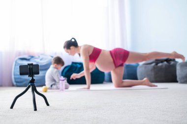Hamile bir kadın oğluyla jimnastik yapıyor. Müstakbel anne telefonda blogu için bir eğitim videosu çekiyor. Bebekli fitness.