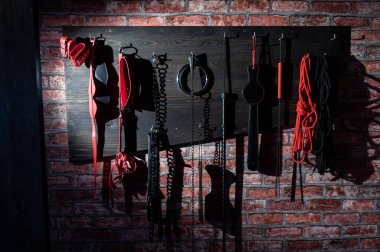 Cinsel zevkler için çeşitli deri kirpikleri tuğla duvardaki ahşap askıda asılı. Rol yapma oyunları için kırmızı ve siyah seks ekipmanları. BDSM odası.