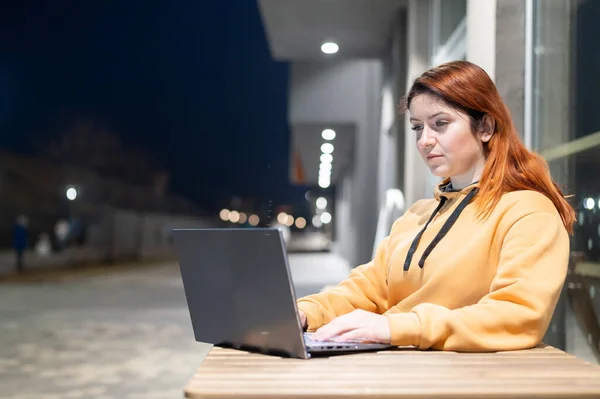 Une femme travaille à distance dans un ordinateur portable dans un café d'été tard dans la soirée. Serious Girl étudiant assis dans une rue vide à une table en bois. Femme pigiste en sweat-shirt . — Photo