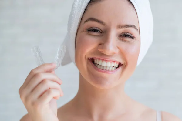 Uma mulher com uma toalha na cabeça segura um protetor bucal clareador para dentes. A menina alinha os dentes com a ajuda de retentores removíveis de noite transparentes. Dispositivo ortodôntico para um sorriso perfeito . — Fotografia de Stock