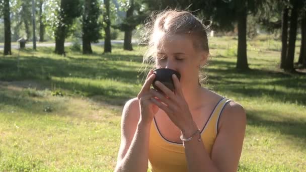 Mujer en una camisa deportiva traga bebida de una taza en el parque . — Vídeos de Stock