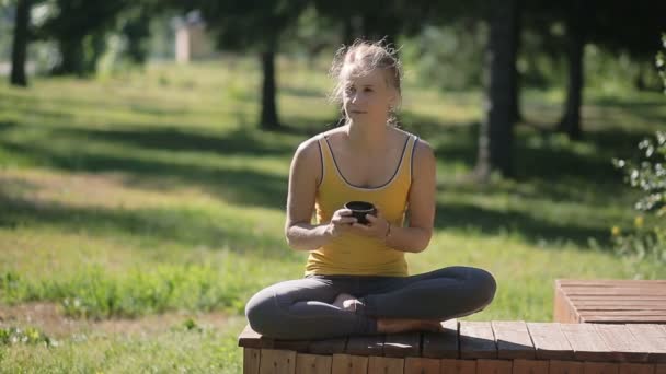 Młoda kobieta siedzi w padmasana pozie i pije herbatę na zewnątrz w lecie. — Wideo stockowe