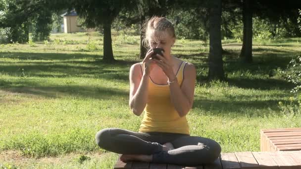 Lachende vrouw zit in lotus pose en dranken drank in de zomer park. — Stockvideo