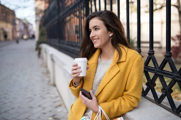 Mujer con un abrigo amarillo bebiendo café de la mañana y usando móvil — Foto de Stock