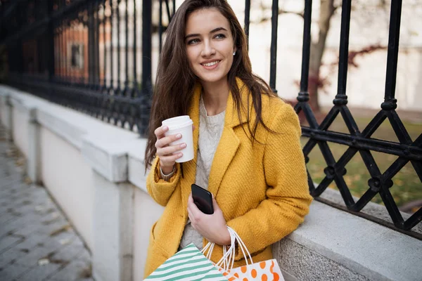 Mujer con un abrigo amarillo bebiendo café de la mañana y usando móvil — Foto de Stock