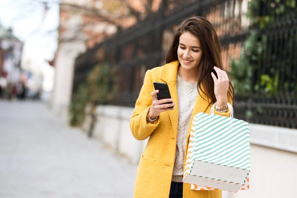 Mujer sonriente en un abrigo amarillo usando móviles y bolsas de espera de las compras — Foto de Stock