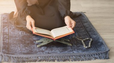 Müslüman bir kadının Kur 'an' dan dua edişinin kırpılmış görüntüsü mavi gri bir hasırın üzerinde, tespih ile birlikte.