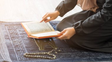 Kesilmiş resim, masadaki Kur 'an' a seçici odaklanma, mavi gri paspasın üzerindeki dua kitabını açan Müslüman kadın ve tespih boncukları.