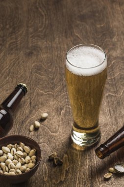 İki koyu bira şişesi, bir bardak bira ve köpük ve ahşap arka planda bir tabak şam fıstığı. - Yakın çekim. Kopyalama. Dikey yönelim.