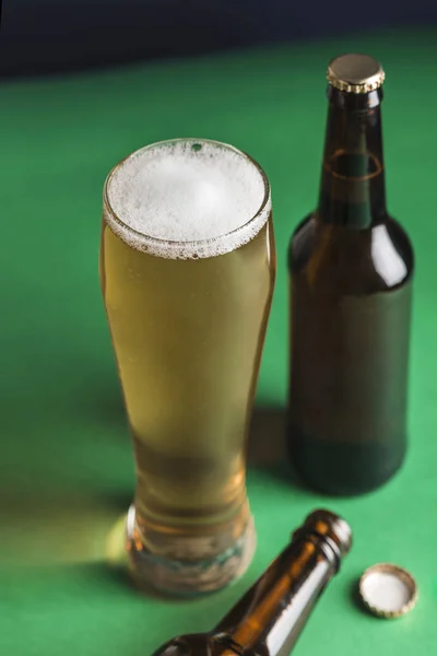 Two dark beer bottles, a glass of beer and foam on a green and blue background. Closeup. Copyspace. Vertical orientation.