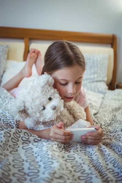 Girl on bed with teddy bear using phone — ストック写真
