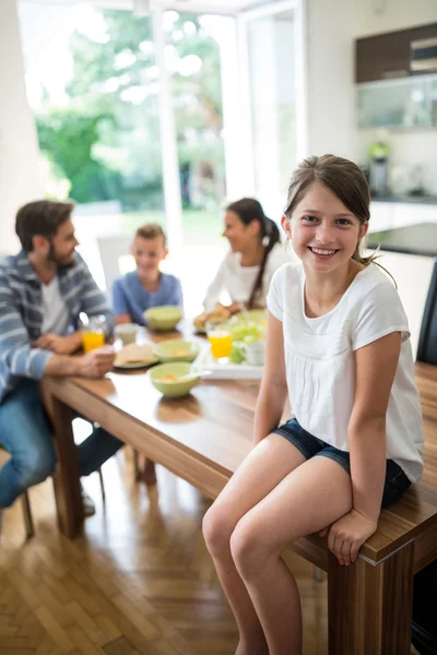 Portrait of girl sitting on dining table while family having breakfast in background — ストック写真