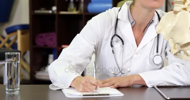 Fisioterapeuta mirando el modelo de columna vertebral mientras escribe en el portapapeles — Vídeos de Stock