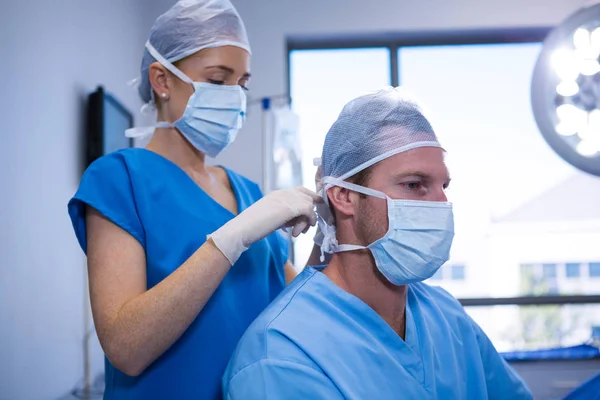 Kvinnlig sjuksköterska hjälper i kopplingsförbehåll kirurgisk mask till hennes medarbetare — Stockfoto