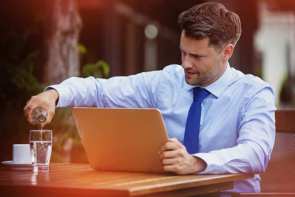 Handsome businessman pouring water in glass while using laptop — Stock Photo, Image