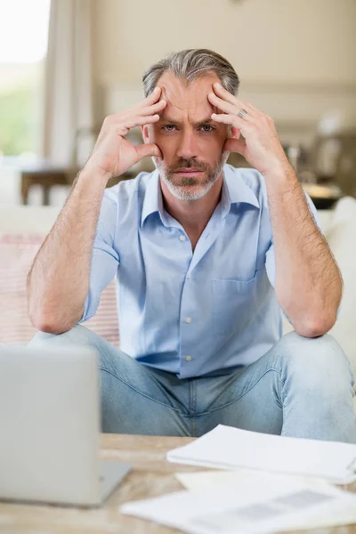 Беспокойный мужчина сидит на диване со счетами и ноутбуком в гостиной — стоковое фото